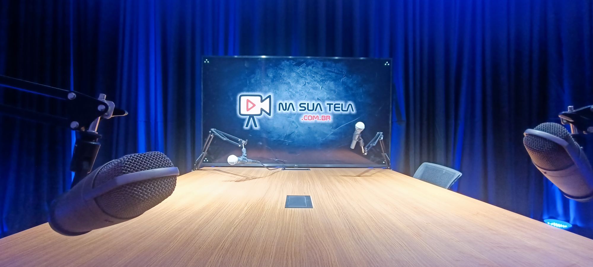 Estúdio NaSuaTela com cortinas azuis, mesa de madeira, microfones de podcast e o logo no telão